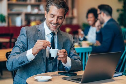 Hombre de unos cuarenta años sonriendo en una cafetería con un portátil y una taza de café, ilustrando un encuentro serio, una primera impresión positiva y una comunicación auténtica.